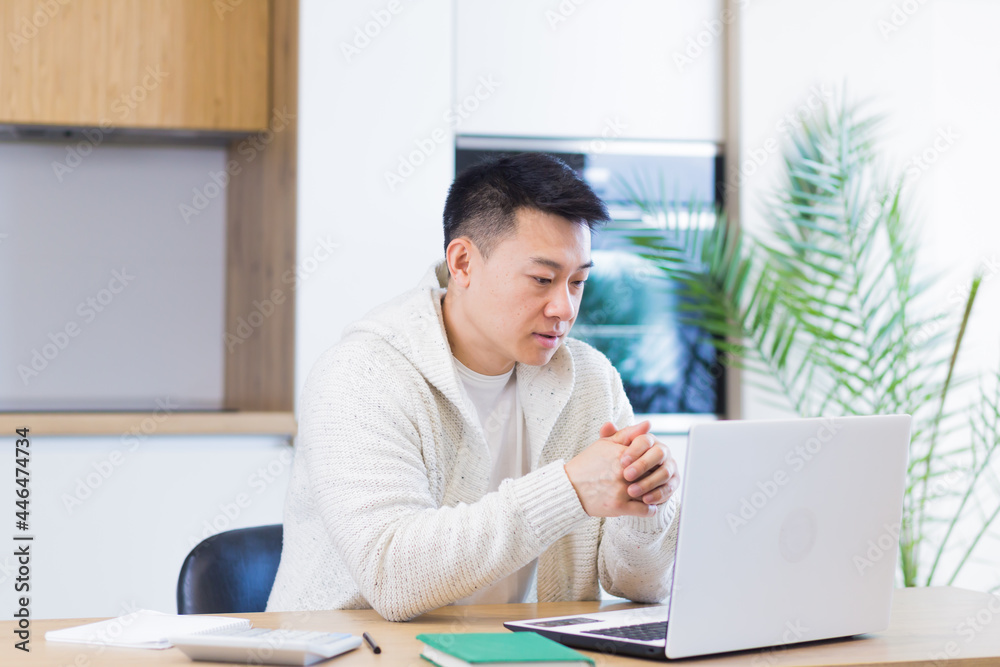 Young asian man working at home on a laptop computer sitting at a table in the kitchen Young asian man working at home on a laptop computer sitting at a table in the kitchen