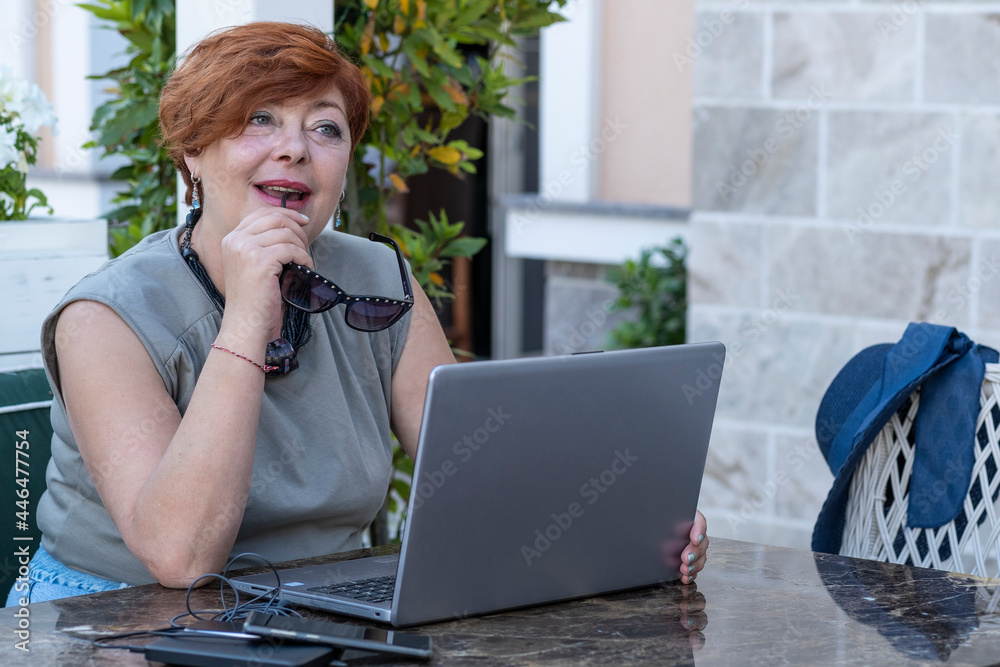 Portrait of a smiling middle-aged woman with short red hair sitting at a table in an open-air cafe holding sunglasses in hand. There is an open laptop and a cup of coffee on the table in front of her