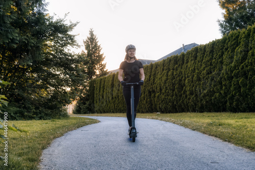 Caucasian Adult Woman riding an electric scooter on a path in a quite neighborhood in a modern city. Taken in Fraser Heights, Surrey, Vancouver, British Columbia, Canada.