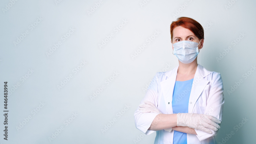 Young woman in white medical gown and protective mask stands with her arms folded on her chest. Portrait of female doctor on gray monochrome background. Banner with medical professional and copyspace