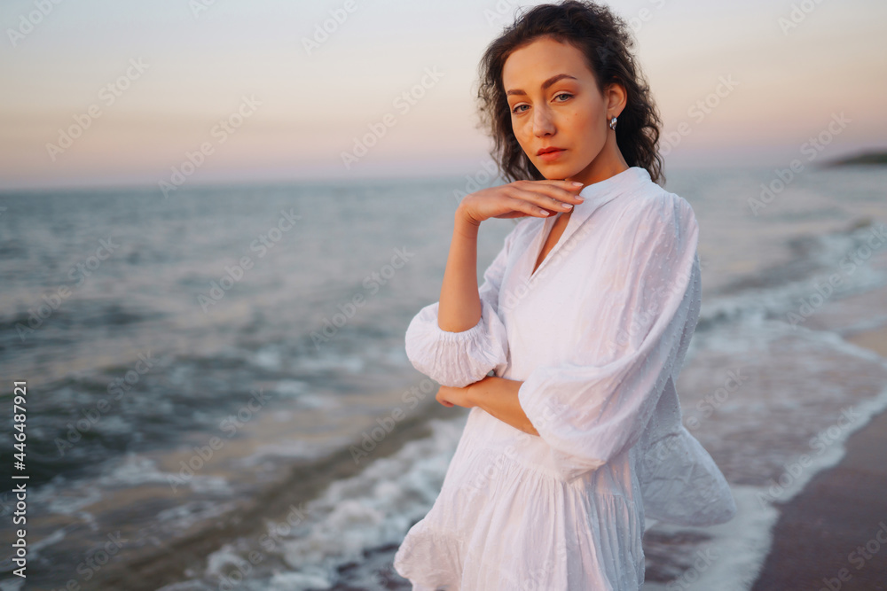 Young happy woman in a white fluttering dress walks along seashore. The girl looks at the magical sunrise. Summer time. Travel, weekend, relax and lifestyle concept. Young happy woman in a white fluttering dress walks along seashore. The girl looks at the magical sunrise. Summer time. Travel, weekend, relax and lifestyle concept.