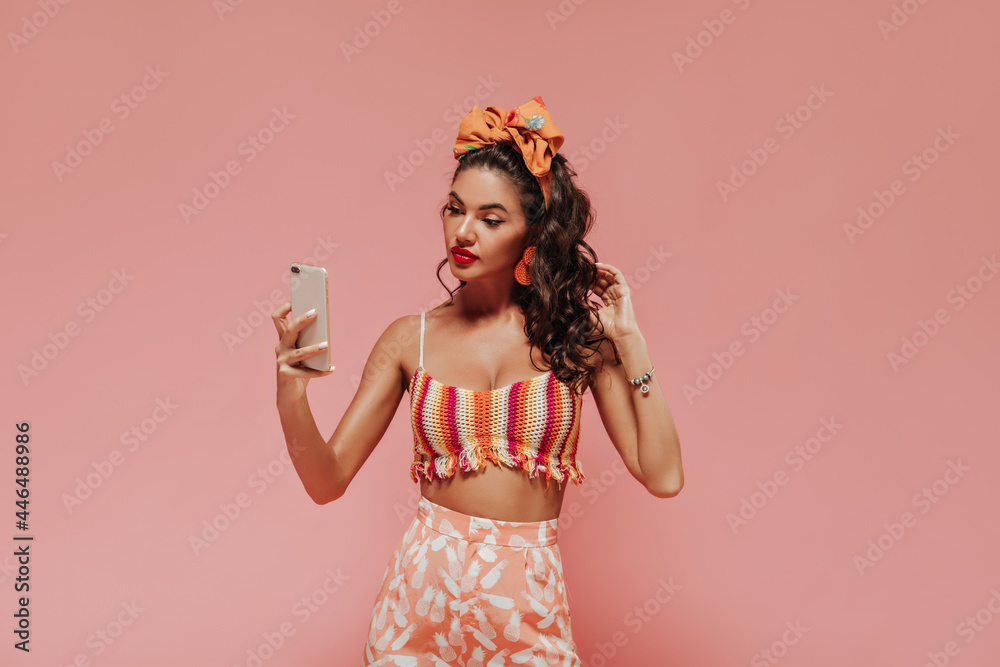 Bright tanned woman with orange bandana and stylish earrings in striped top and printed pink pants makes selfie on isolated backdrop.. Bright tanned woman with orange bandana and stylish earrings in striped top and printed pink pants makes selfie on isolated backdrop..