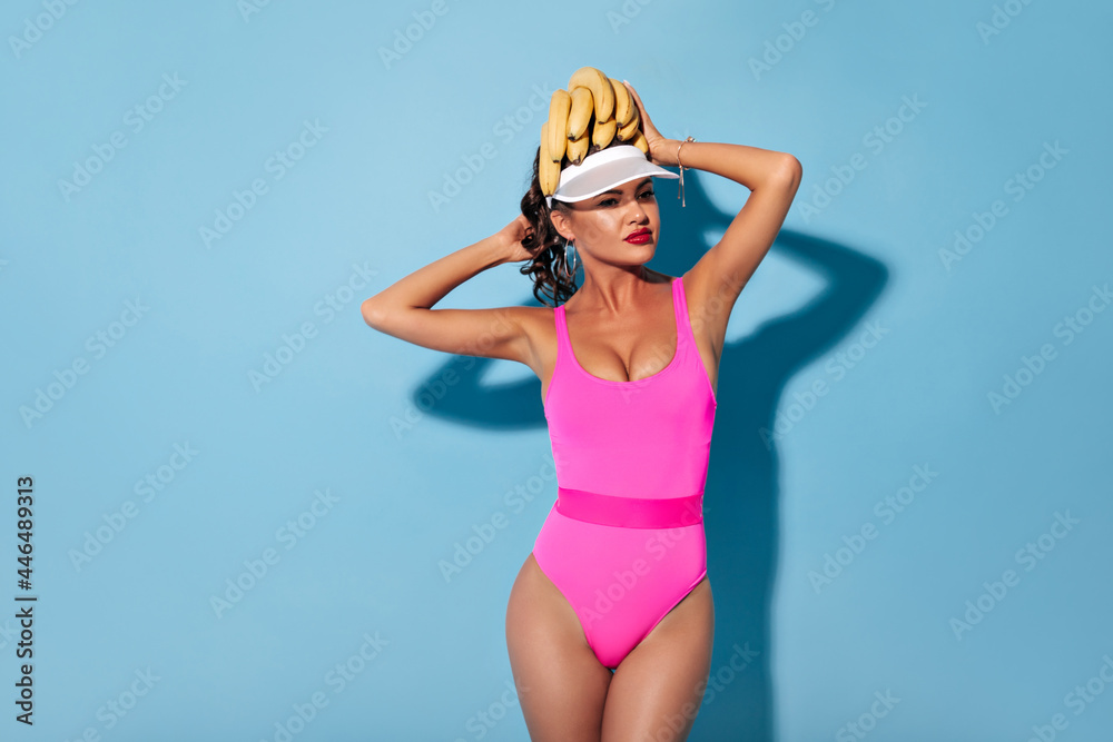Wonderful bright young woman with red lips and curly hair in summer pink clothes holding bananas on her head and looking away..