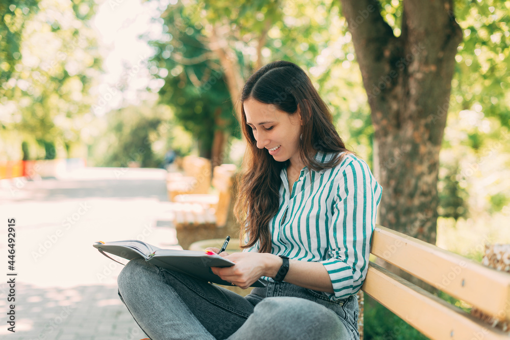 Fototapeta premium Photo of smiling young woman sitting on bench with pen and book, in park and writing in planner