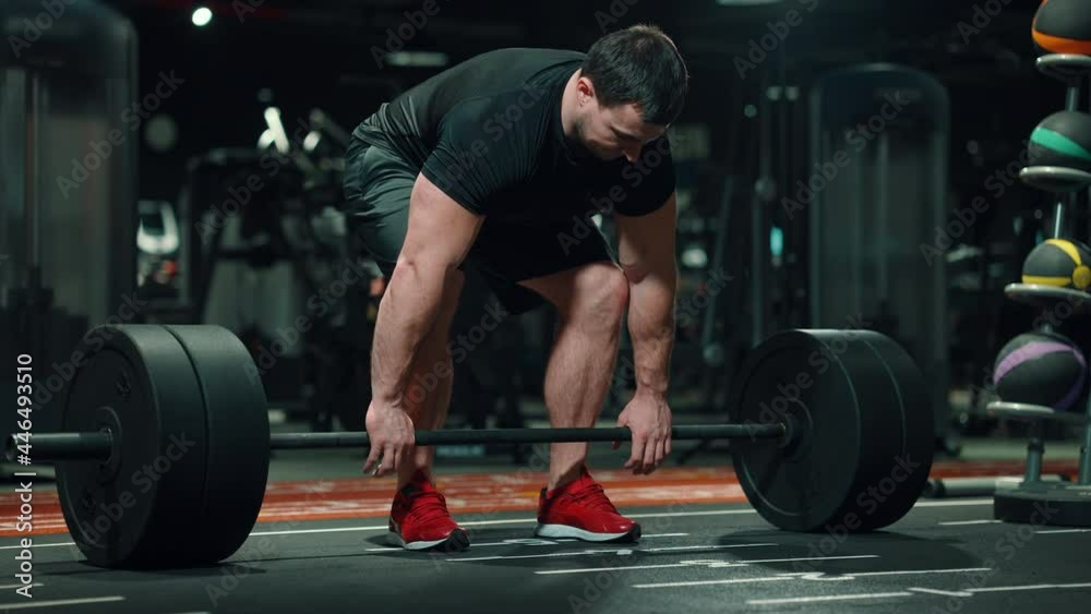 Muscular man wearing black sportswear lifting barbell in gym, equipment on blurred background. Low shot of bodybuilder working out. Concept of sport Muscular man wearing black sportswear lifting barbell in gym, equipment on blurred background. Low shot of bodybuilder working out. Concept of sport