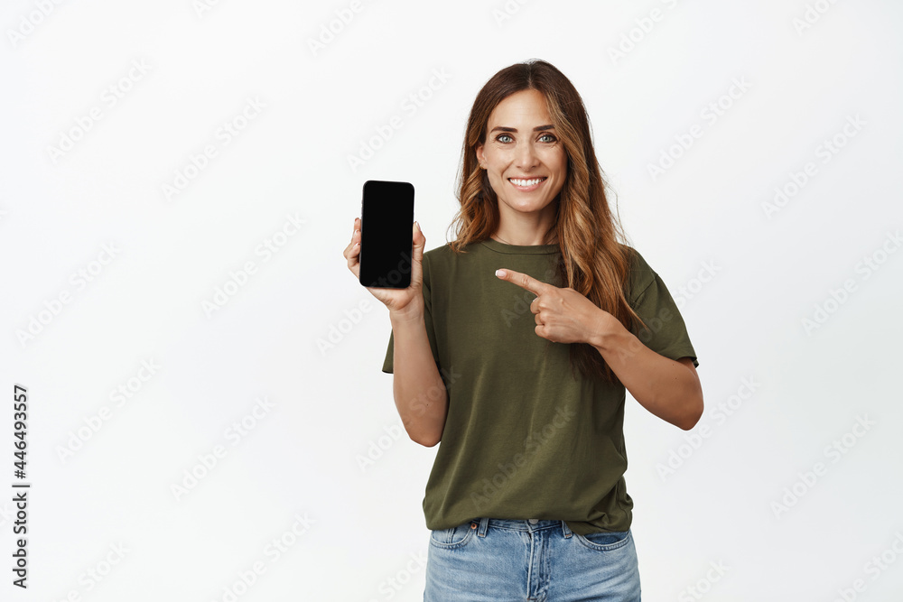 Fototapeta premium Portrait of smiling adult woman pointing finger at screen of mobile phone, showing interface, recommend application, website of store or company, new feature in app, white background