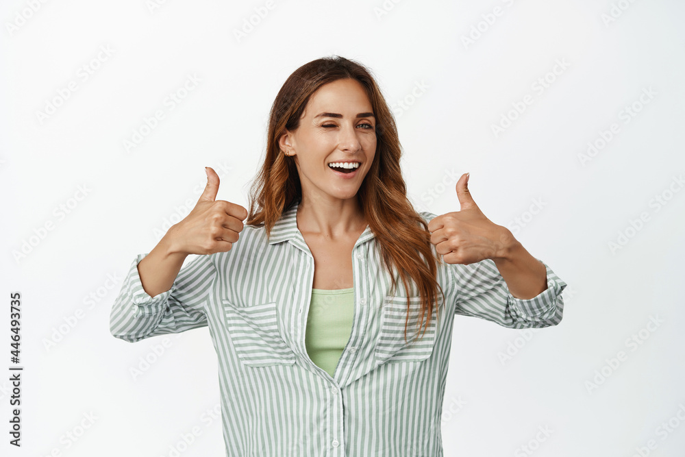 Portrait of happy smiling woman winking, showing thumbs up in approval, like and agree, praise smth good, complimenting you, hinting on smth, standing over white background Portrait of happy smiling woman winking, showing thumbs up in approval, like and agree, praise smth good, complimenting you, hinting on smth, standing over white background