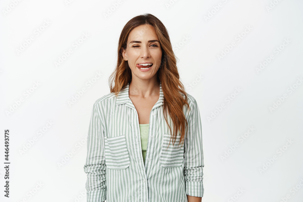 Cheerful brunette woman winking and showing tongue, having fun, looking happy, standing in blouse against white background Cheerful brunette woman winking and showing tongue, having fun, looking happy, standing in blouse against white background