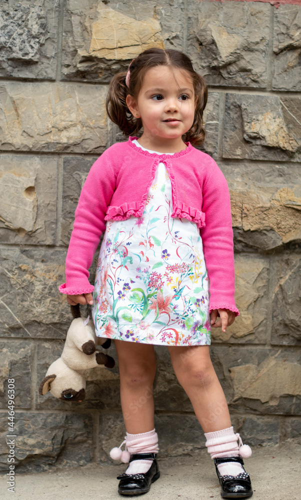 Niña con vestido de flore y chaqueta rosa jugando jovial con un peluche de perrito.
