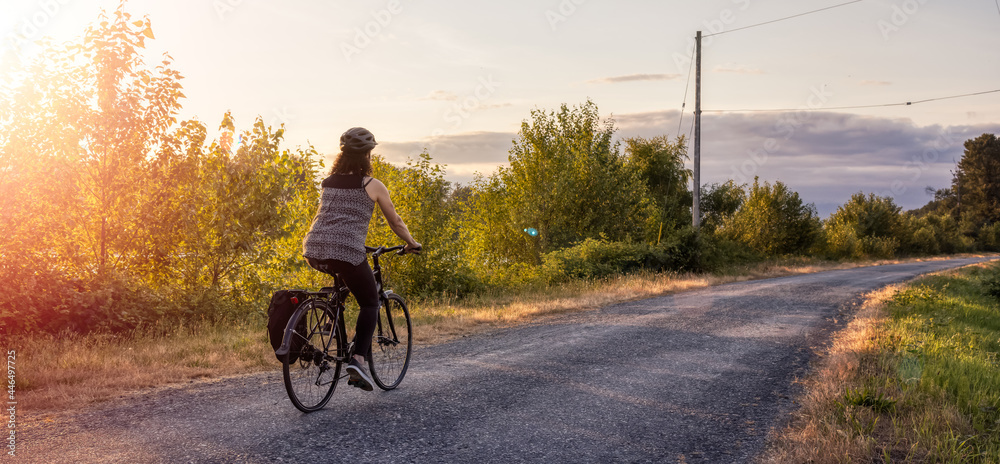 Adventurous White Cacasusian Woman riding a bicycle on a road. Sunny Summer Sunset. Barnston Island, Vancouver, British Columbia, Canada. Adventure Journey Concept