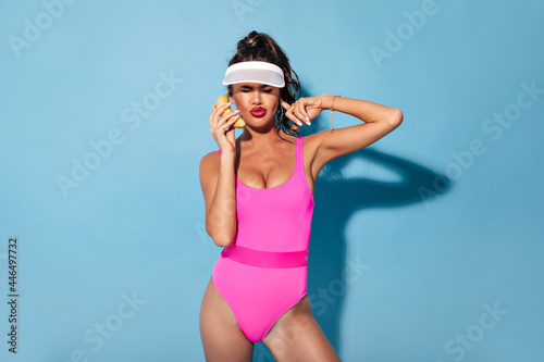 Charming fashionable girl with stylish hairstyle and round earrings in cool swimming suit and cap posing with banana on isolated blackdrop.. © Look!