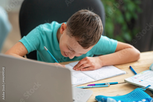 inquisitive boy uses laptop to study school subjects during his online lesson at home. Writing down topic lesson in notebook. E-Education Distance Home Education.