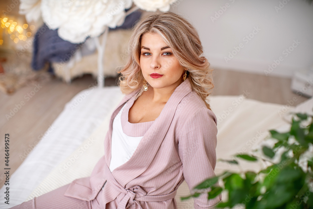 Naklejka premium Portrait of an attractive beautiful young woman in an elegant outfit in a bright interior. a pretty girl with curly blonde hair, in a beautiful dress, with beautiful makeup and styling looks away.