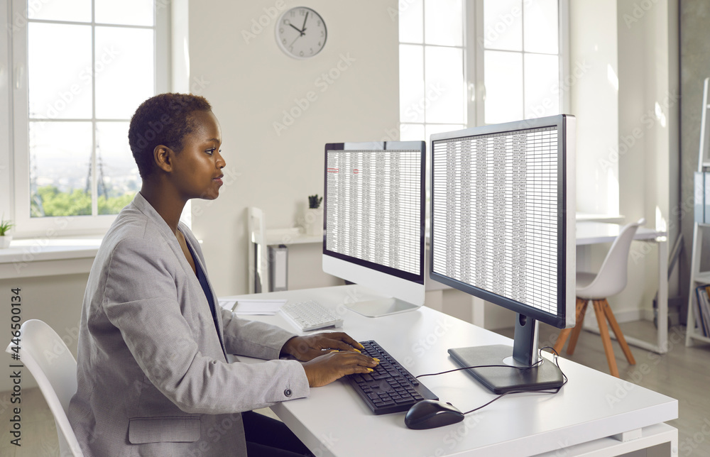 Black business lady or financial accountant using modern bookkeeping software. Serious young woman in suit sitting at office desk and working with data lists on two desktop computers - obrazy, fototapety, plakaty 