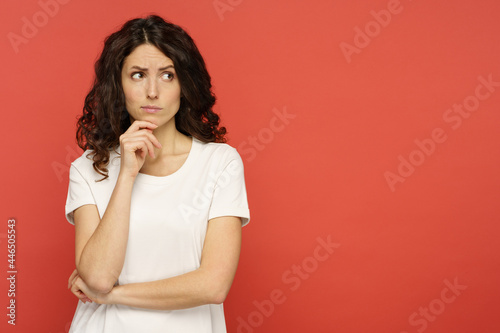 Doubtful young woman touching her chin, skeptic and pensive think with folded arms looking up to copy space over red studio background. Confident serious girl ponder on question or solution photo