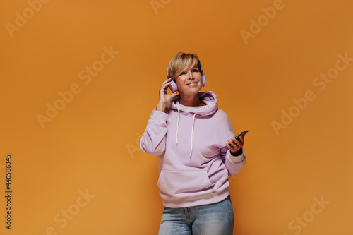 Smiling trendy old lady with blonde cool hairstyle in pink sweatshirt and light jeans posing with lilac headphones and smartphones..