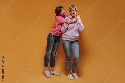 Short brunette haired young lady in pink fashionable sweatshirt smiling and hugging old trendy woman in lilac hoodie on orange backdrop.. © Look!