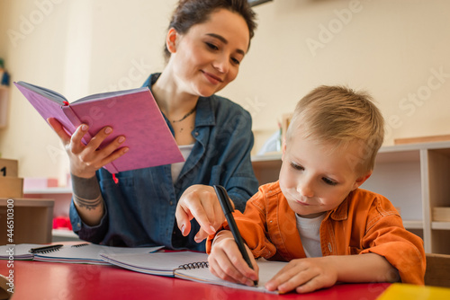 Фотография smiling teacher pointing with finger near kid writing dictation in montessori sc