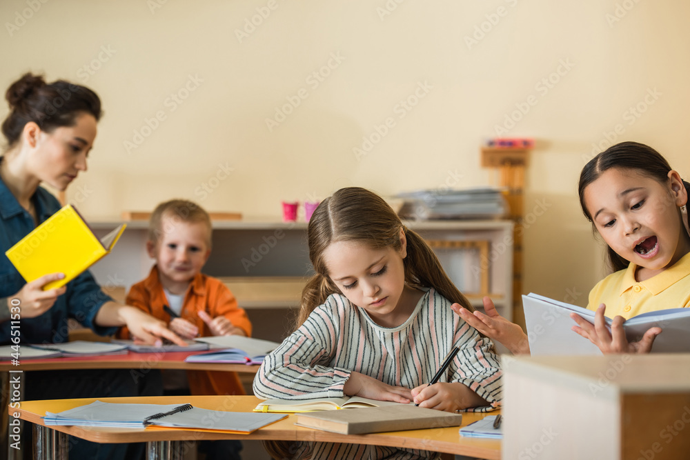 asian girl reading book near blurred teacher and children writing in notebooks during lesson asian girl reading book near blurred teacher and children writing in notebooks during lesson