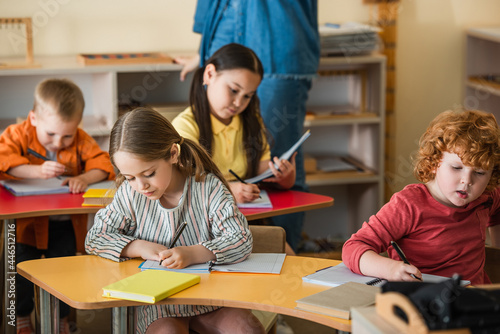 multiethnic children writing in notebooks near teacher on blurred background