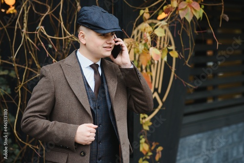 A man posing in the image of an English retro gangster of the 1920s dressed in Peaky blinders style near old brick wall.