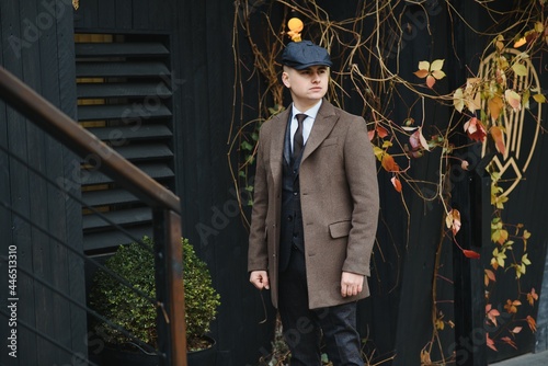 A man posing in the image of an English retro gangster of the 1920s dressed in a coat, suit and flat cap in Peaky blinders style.