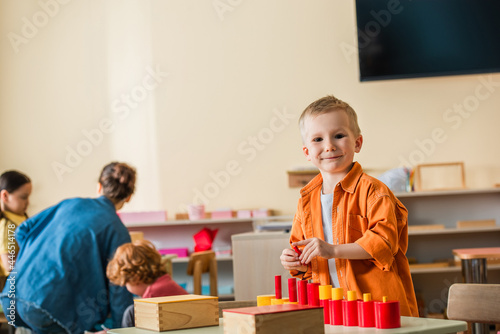 cheerful boy looking at camera near tower made from red cylinders and blurred teacher with interracial girls