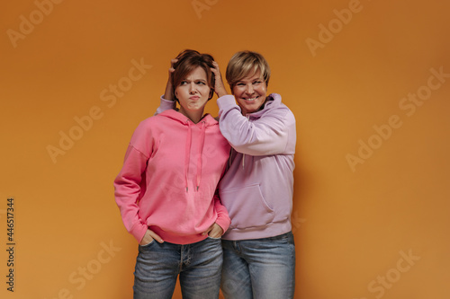 Joyful blonde haired woman in lilac hoodie smiling and posing with sad young girl in pink clothes on orange isolated backdrop..