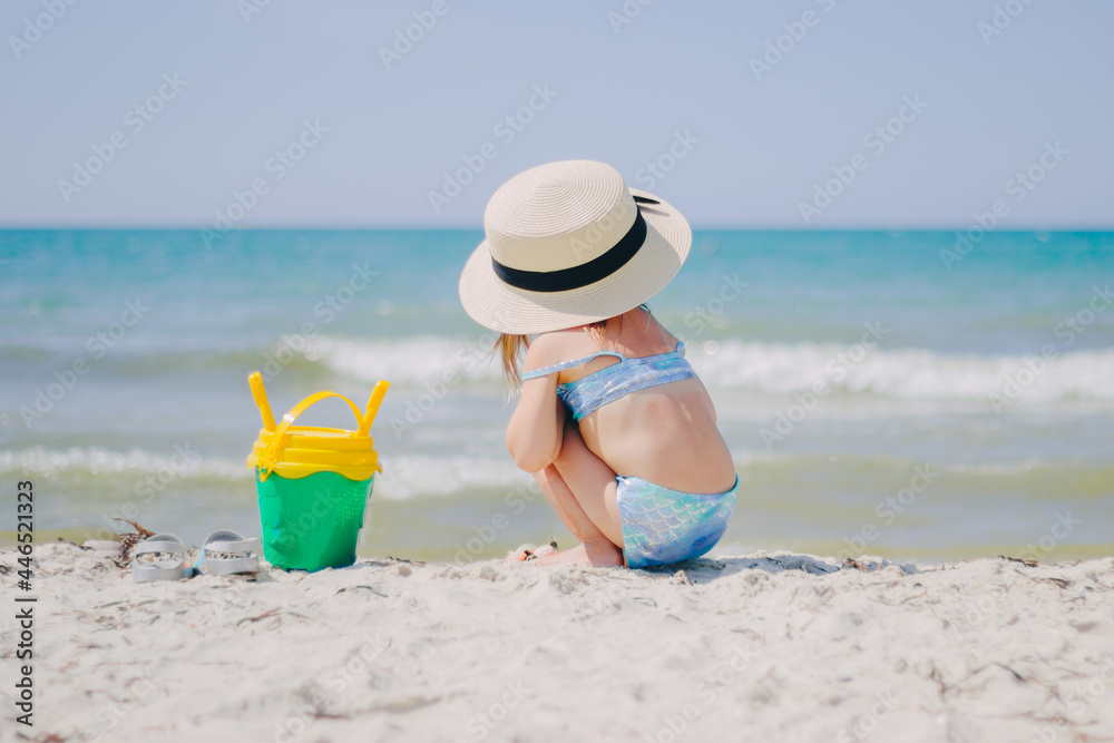 Little girl in a blue mermaid swimsuit sits backwards on the seashore in a straw hat and with a bucket of sand set for playing Little girl in a blue mermaid swimsuit sits backwards on the seashore in a straw hat and with a bucket of sand set for playing
