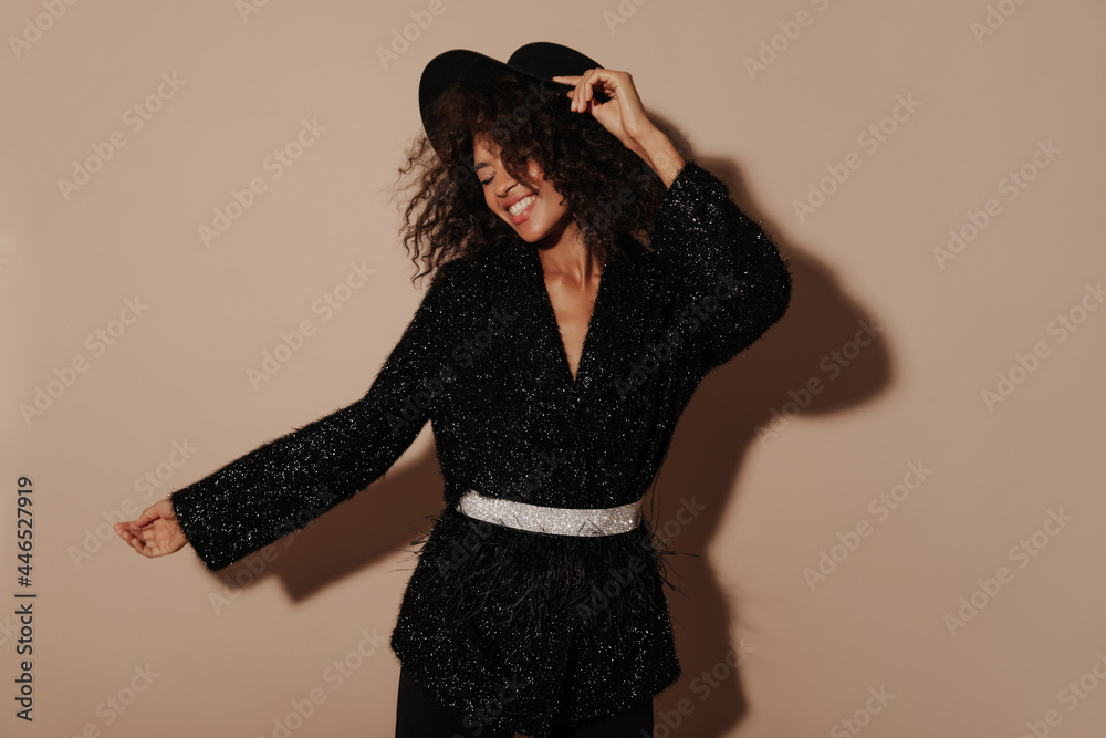 Positive curly haired girl with modern hat and shiny black long sleeve sweater with belt smiling with closed eyes on beige backdrop.. Positive curly haired girl with modern hat and shiny black long sleeve sweater with belt smiling with closed eyes on beige backdrop..