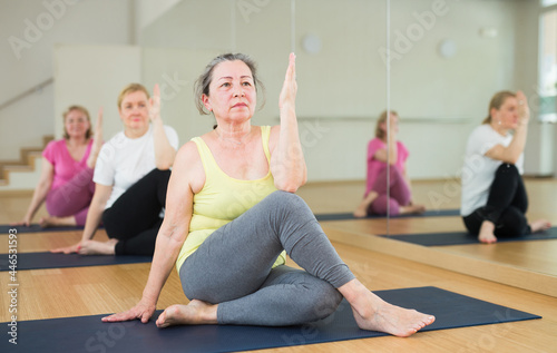 Active elderly woman practicing twisting yoga postures on mat at group class