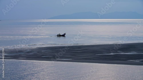 Static wide shot of Longtail boat in silhouette with local fishermen sailing through calm water at sunset in Thailand. Sand bank cuts through the foreground with blue karst mountains in background. photo