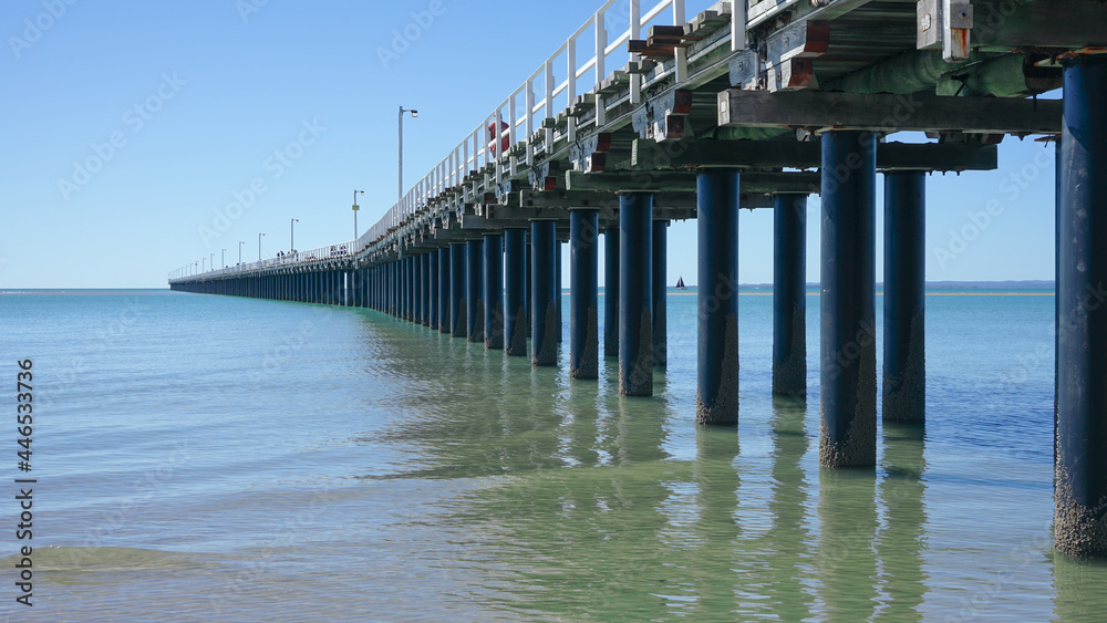 Perspective view of the Urangan Pier from the shoreline with gentle waves, blue sky and a distant sailing boat. 