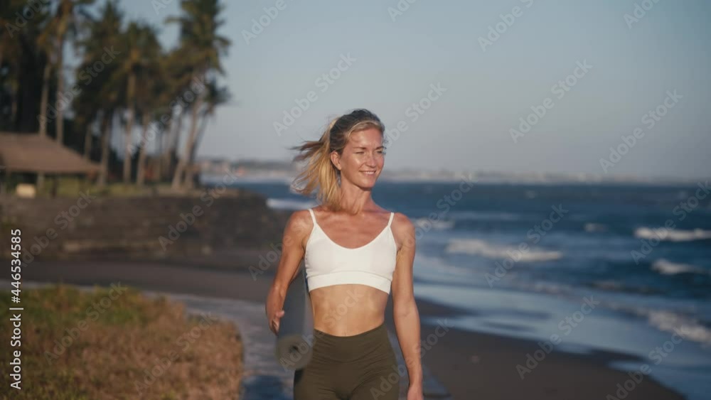 Attractive woman holding yoga mat walking along shore breathing fresh air, Bali Attractive woman holding yoga mat walking along shore breathing fresh air, Bali