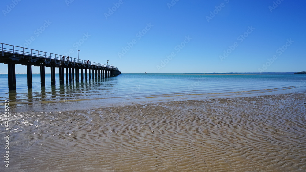 View of the bay with rippled sand, gentle waves, blue sky and a jetty reaching into the distance