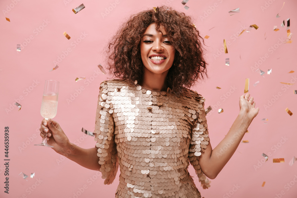 Positive brunette woman in modern beige dress smiling and posing with glass with wine and with confetti on pink backdrop.. Positive brunette woman in modern beige dress smiling and posing with glass with wine and with confetti on pink backdrop..