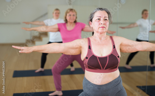 Three mature European women are doing warrior II pose in fitness class
