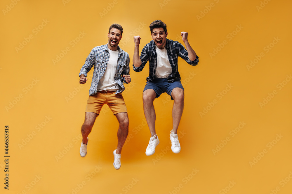 Young active optimistic men in checkered shirts and colorful shorts jump high and rejoice on isolated orange background.