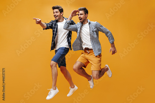 Cheerful young happy men in colorful shorts and checkered blue shirts point into distance, smile and jump on orange background.
