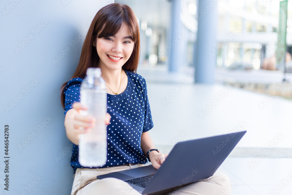 Happy young attractive Asian woman smiling and holding out the bottle of water to the camera while working on her laptop computer Happy young attractive Asian woman smiling and holding out the bottle of water to the camera while working on her laptop computer