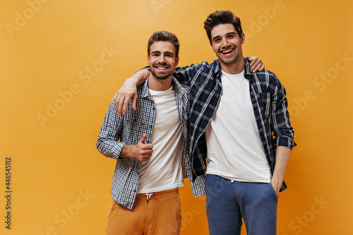 Charming men in checkered shirts and white t-shirt smile on orange background. Cool bearded guy shows peace sign. photo