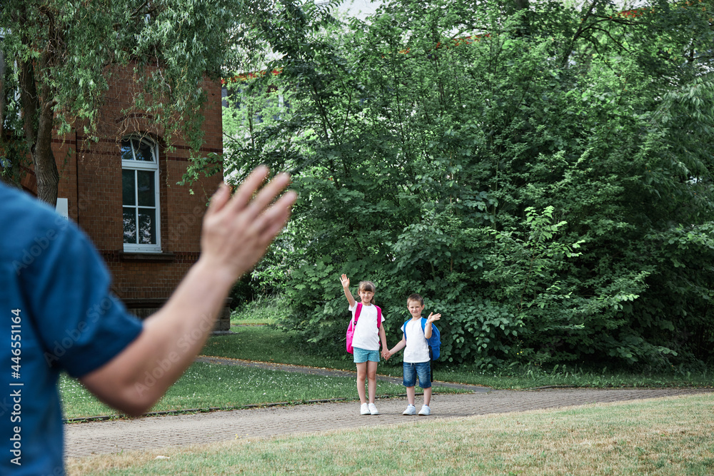 Obraz premium Dad takes the kids to school or kindergarten, stands and waves at them. Happy kids return to school after vacation. Morning rituals, school days