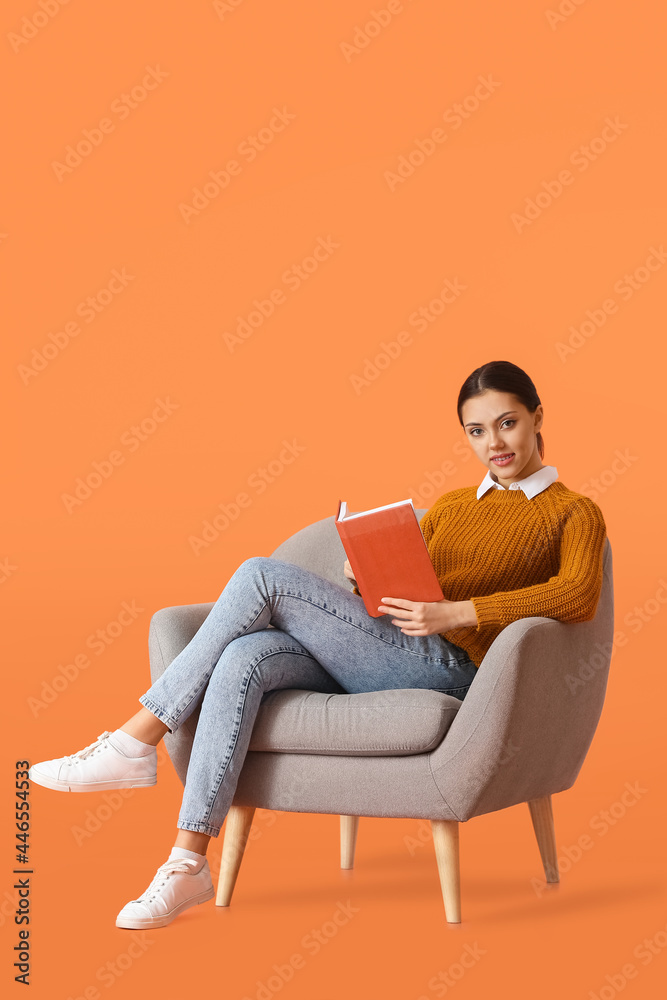 Beautiful young woman with book sitting in armchair on color background