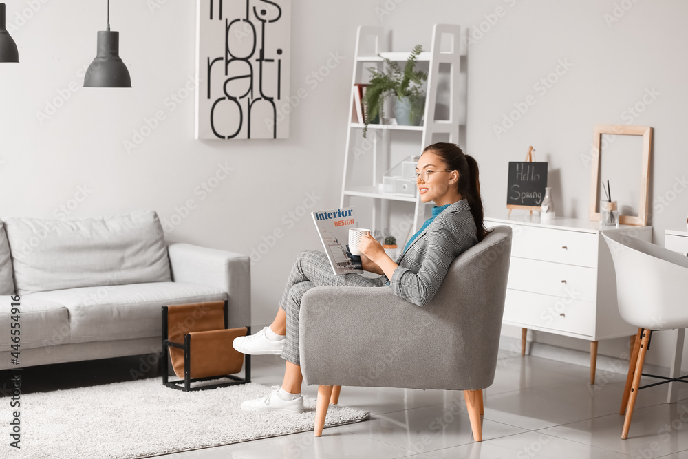 Fototapeta premium Young woman with coffee and magazine sitting in armchair at home