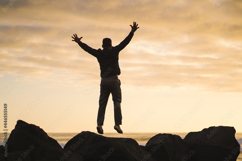 happy jumping MAN  in YES POSE on beach rocks AT THE ocean WITH arms wide open enjoying life
