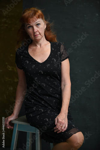 Vertical portrait of a caucasian adult red-haired woman sitting on a chair against a dark background looking at the camera