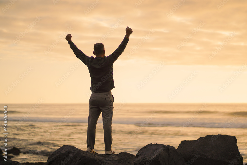 happy successful MAN  in YES POSE on beach rocks AT THE ocean WITH arms wide open enjoying life
