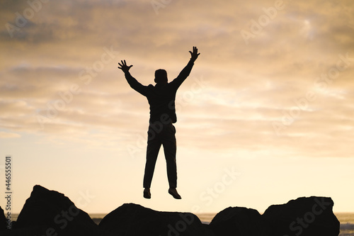happy jumping MAN in YES POSE on beach rocks AT THE ocean WITH arms wide open enjoying life 