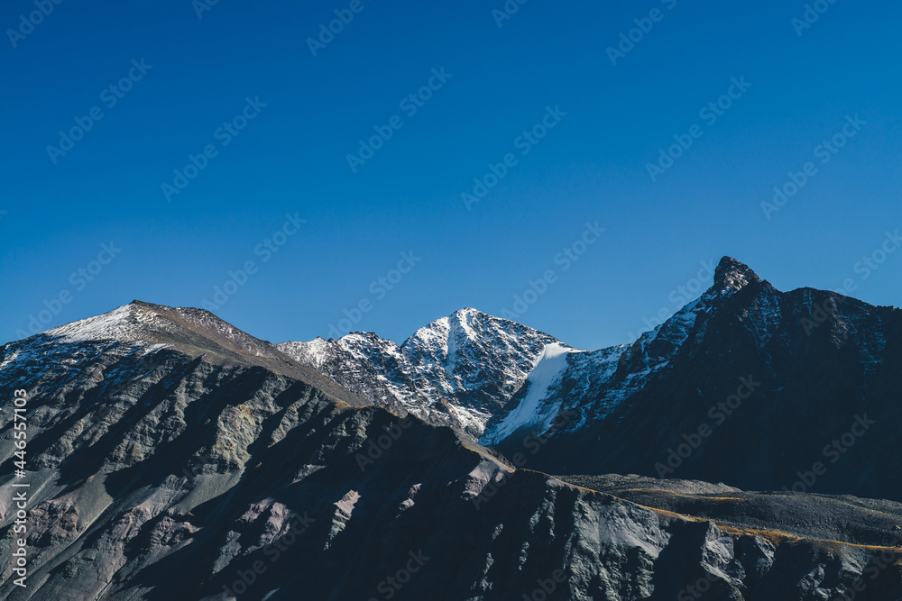 Atmospheric mountain landscape with dark mountains silhouettes. Sharp rocky pinnacle and snow-covered top in sunlight. Alpine scenery with black orange mountain with peaked top in sunshine in autumn.