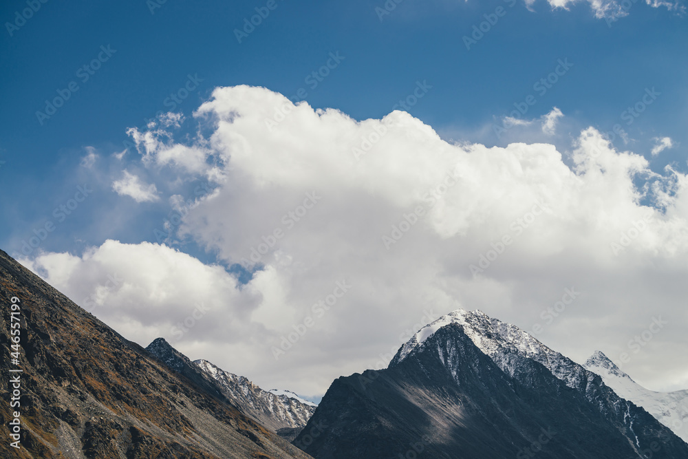 Atmospheric alpine landscape with high mountain silhouette with snow on peaked top under blue cloudy sky. Dramatic mountain scenery with beautiful snow-covered pointy peak and high snowy mountain wall